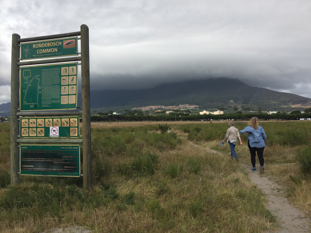 Two women walking on a dirt path in grass, near a sign