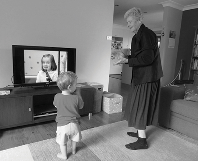 Toddler and older woman standing in front of a TV looking at each other and clapping their hands.