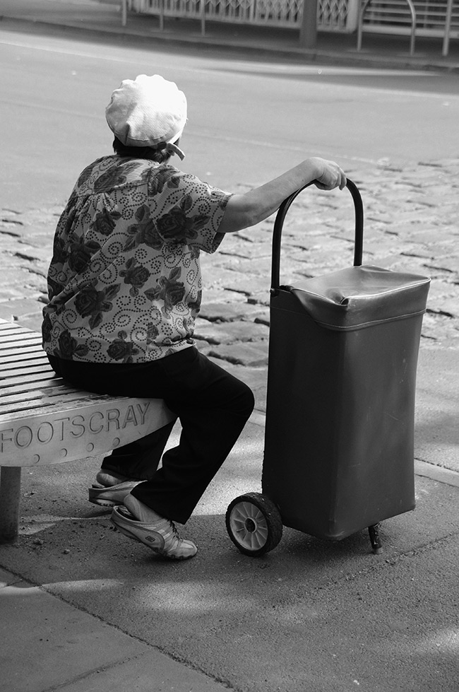 Woman with shopping trolley sitting on bench that reads Footscray